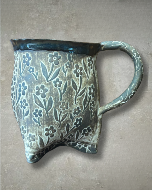 A handmade ceramic mug with an uneven base and a curved handle, featuring a carved floral pattern and earthy tones of green and brown.