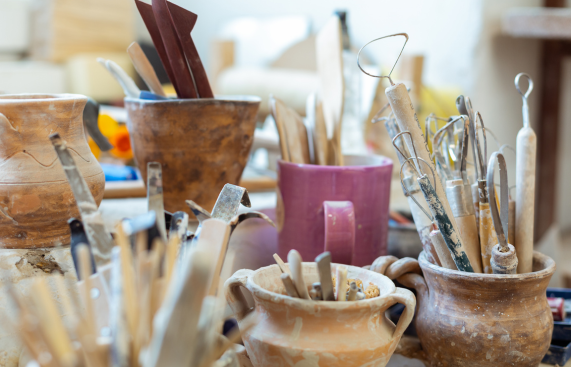 A variety of pottery tools such as loop tools, carving tools, and ribs organized in clay pots and a mug on a cluttered workbench in a ceramics studio.