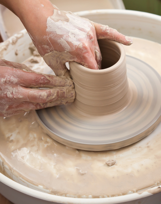 Close-up of hands shaping a clay vessel on a spinning pottery wheel, covered in wet clay during the wheel-throwing process.